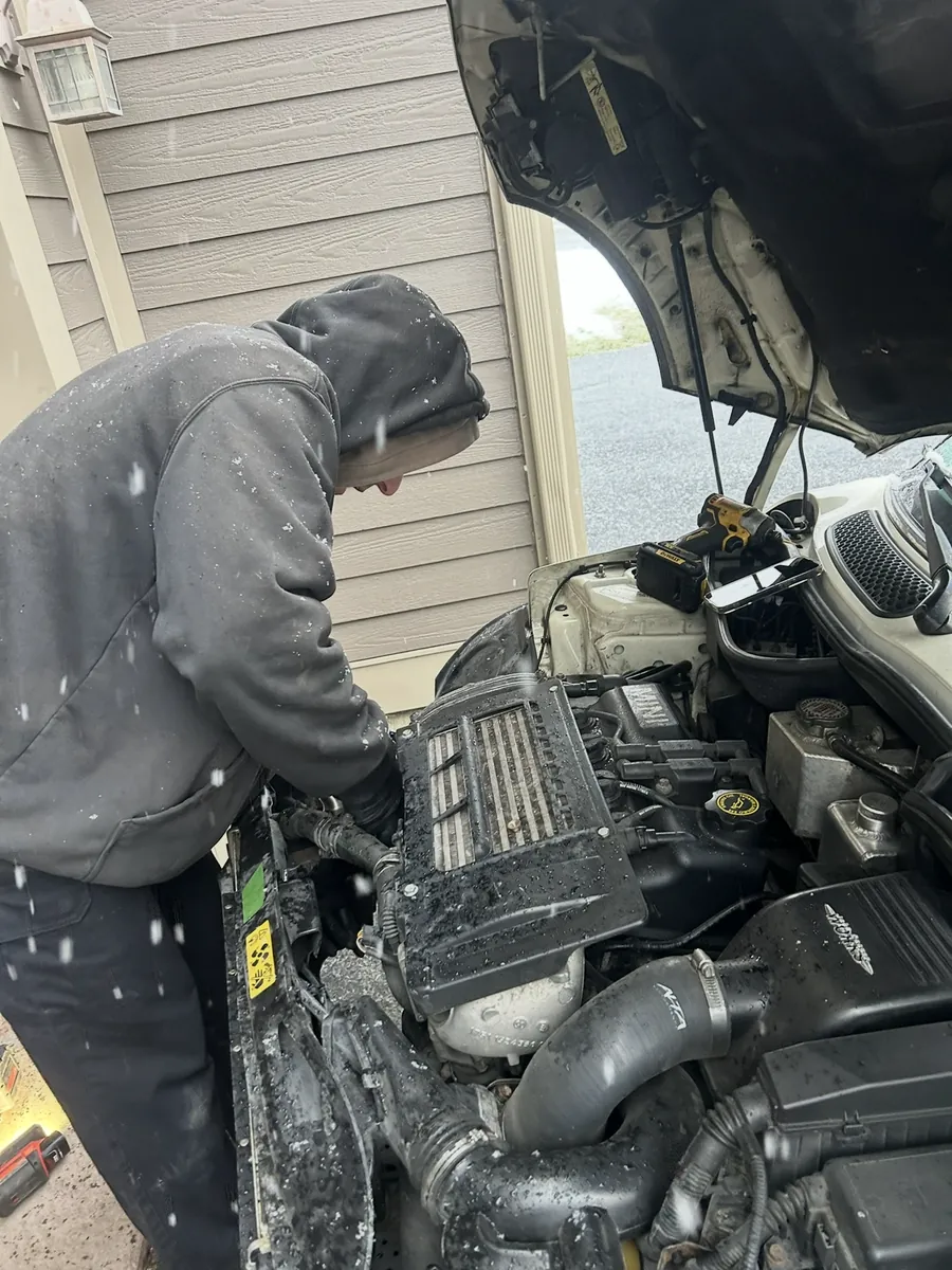 Working under the hood of a car in a driveway as snow falls